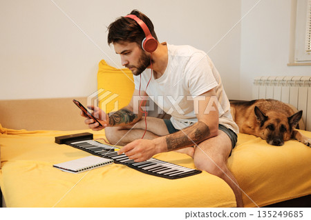 A man sits on a bed with headphones and checks his phone while working on a roll-up keyboard. The scene highlights home music creation and the concept of digital assistance 135249685