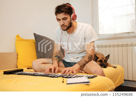 A man sits on a bed with headphones on and plays a roll-up keyboard while a dog lies beside him. The scene shows concentrated home music practice and the concept of creative work 135249686