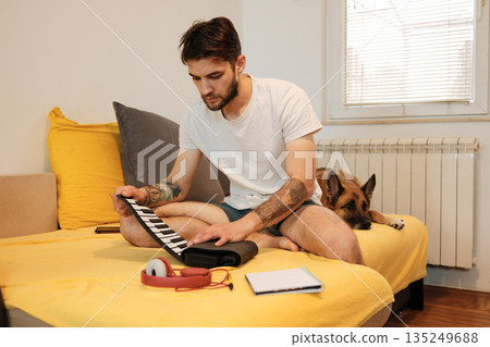A man sits on a bed and practices on a small roll-up keyboard while a dog rests nearby. The scene shows a quiet creative moment and the concept of home music work 135249688