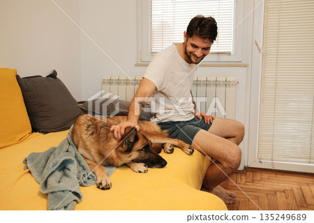 A man sits on a bed and gently pets a German Shepherd, capturing a calm morning moment and human pet connection A man sits on a bed and gently pets a German Shepherd, capturing a calm morning moment and human pet connection 135249689