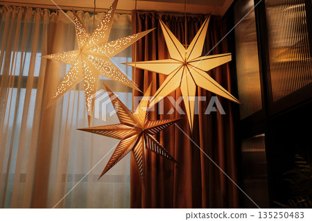 Three decorative star-shaped lamps glowing warmly against a curtain backdrop in a cozy room. The concept of minimalistic Christmas decor Three decorative star-shaped lamps glowing warmly against a curtain backdrop in a cozy room. The concept of minimalistic Christmas decor 135250483
