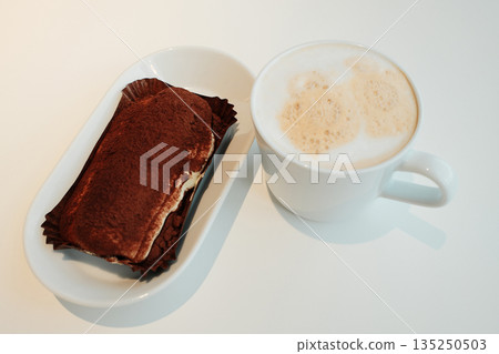 Classic tiramisu topped with cocoa powder served beside a foamy cappuccino on a white table, minimalist cafe setting. The concept of breakfast in a cafe Classic tiramisu topped with cocoa powder served beside a foamy cappuccino on a white table, minimalist cafe setting. The concept of breakfast in a cafe 135250503