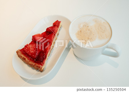 Slice of glazed strawberry tart with golden crust served beside a cup of cappuccino on a clean white surface. The concept of breakfast in a cafe 135250504