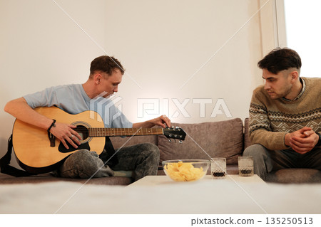 A man tunes his guitar while his friend watches him from the couch. The concept emphasizes creativity, focus, and relaxed artistic moments at home 135250513