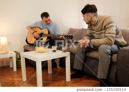 A man plays guitar on the couch while his friend listens attentively. The concept conveys creativity, expression, and shared appreciation of music A man plays guitar on the couch while his friend listens attentively. The concept conveys creativity, expression, and shared appreciation of music 135250514