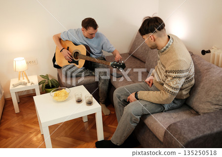 A man tunes his guitar while his friend watches him from the couch. The concept emphasizes creativity, focus, and relaxed artistic moments at home A man tunes his guitar while his friend watches him from the couch. The concept emphasizes creativity, focus, and relaxed artistic moments at home 135250518