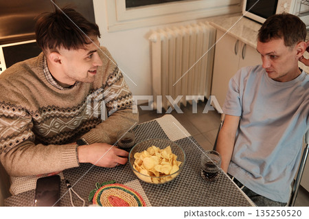 Two friends sit at a kitchen table with snacks and drinks, talking casually. The concept focuses on connection, simple social time, and authentic everyday living Two friends sit at a kitchen table with snacks and drinks, talking casually. The concept focuses on connection, simple social time, and authentic everyday living 135250520