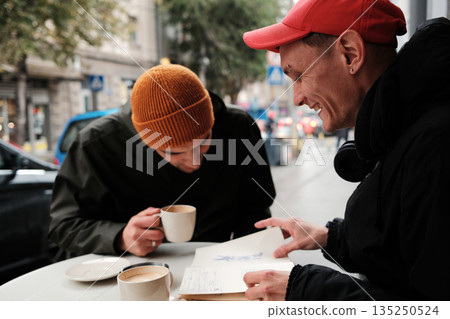 Two men review hand drawn sketches together at an outdoor table. The scene highlights creativity concept and collaboration concept. Two men review hand drawn sketches together at an outdoor table. The scene highlights creativity concept and collaboration concept. 135250524