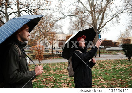 A man under an umbrella takes a photo with his smartphone in a rainy autumn park. The scene represents the concept of creativity and personal expression A man under an umbrella takes a photo with his smartphone in a rainy autumn park. The scene represents the concept of creativity and personal expression 135250531