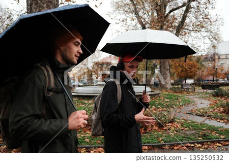 Two men walk under umbrellas while talking in a rainy city park. The scene reflects the concept of communication and shared time Two men walk under umbrellas while talking in a rainy city park. The scene reflects the concept of communication and shared time 135250532