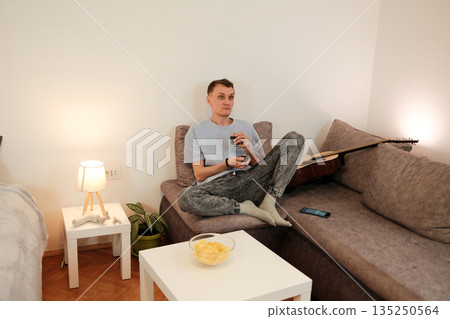 A young man sits on a sofa with a drink in hand and watches television attentively. The moment represents the concept of relaxation and evening routine 135250564