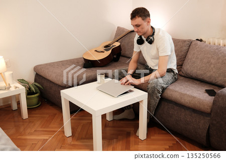 A man sits on a sofa and closes his laptop after finishing his tasks. The scene conveys the concept of work routine and transition to rest A man sits on a sofa and closes his laptop after finishing his tasks. The scene conveys the concept of work routine and transition to rest 135250566