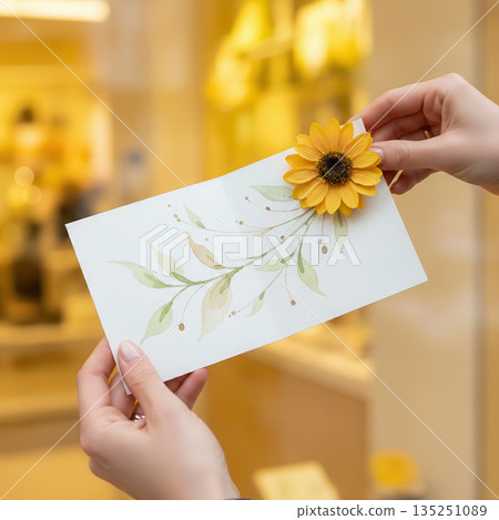 Hands holding a decorative card featuring a sunflower and botanical illustration, showcasing creativity and artistry in a bright, inviting environment with soft lighting Hands holding a decorative card featuring a sunflower and botanical illustration, showcasing creativity and artistry in a bright, inviting environment with soft lighting 135251089