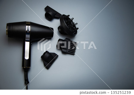 The black hair dryer lies on the side and next to it there is a diffuser nozzle, a comb nozzle and classic nozzles of different diameters on a gray background, a top view with space for text. The 135251239