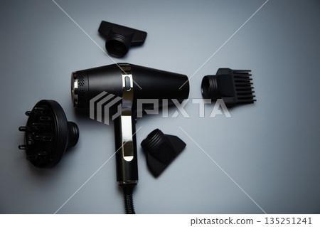 A black hair dryer around which there are different drying attachments - a comb, a diffuser and classic nozzles of different diameters on a gray background, top view. High quality photo A black hair dryer around which there are different drying attachments - a comb, a diffuser and classic nozzles of different diameters on a gray background, top view. High quality photo 135251241