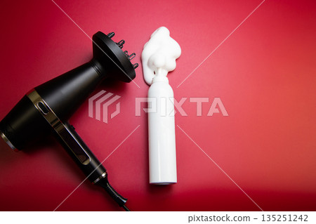 A top view of the foam for styling and fixing the hairstyle in a white bottle, next to it is a hairdryer with a nozzle - a diffuser for a hairdryer on a pink background with space for text. The A top view of the foam for styling and fixing the hairstyle in a white bottle, next to it is a hairdryer with a nozzle - a diffuser for a hairdryer on a pink background with space for text. The 135251242