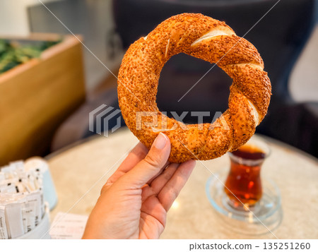 Hand holding traditional Turkish simit with tea in background. Culture, nutrition, and lifestyle in everyday travel and hospitality settings. 135251260