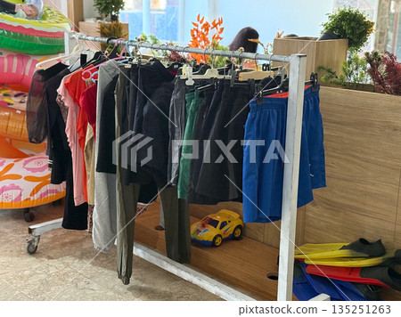 Clothes hanging on a rack with toys and inflatables in the background of a store. Retail, lifestyle and vacation essentials reflecting shopping, leisure and summer atmosphere. 135251263