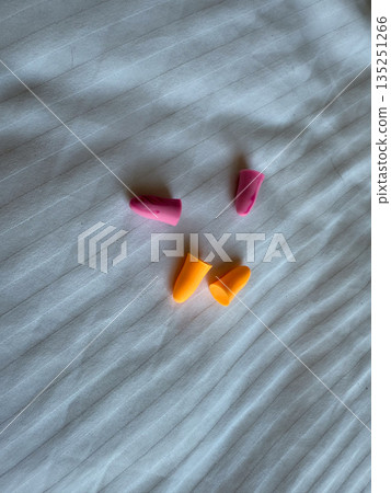 Foam earplugs in pink and orange color lying on a white bed sheet. Health, sleep, protection, lifestyle accessory and travel accommodation detail. Foam earplugs in pink and orange color lying on a white bed sheet. Health, sleep, protection, lifestyle accessory and travel accommodation detail. 135251266