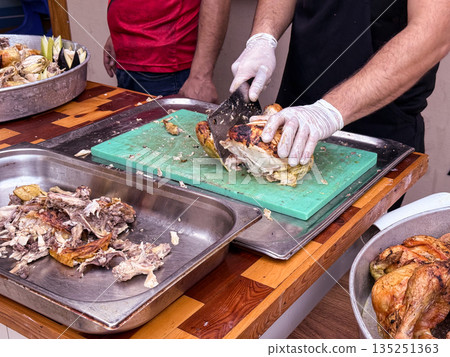 Cook slicing roasted chicken on a cutting board in a kitchen setting. Culinary preparation, food service, and gastronomy. Cook slicing roasted chicken on a cutting board in a kitchen setting. Culinary preparation, food service, and gastronomy. 135251363