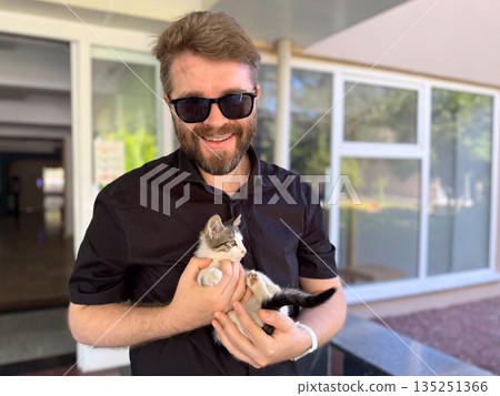 Man in sunglasses smiling and holding small kitten outdoors. Companionship affection, and emotional bond between human and animal. 135251366