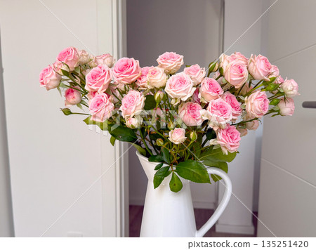 Close-up bouquet of pink roses in white ceramic jug placed on minimal white table indoors. Floral arrangement, interior decor, and lifestyle aesthetic in a modern home setting. 135251420