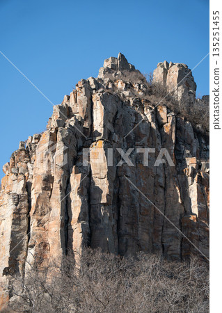 Dramatic rocky cliff rises against a clear blue sky in Armenia, vertical stone formations, warm earth tones, rugged mountain landscape Dramatic rocky cliff rises against a clear blue sky in Armenia, vertical stone formations, warm earth tones, rugged mountain landscape 135251455
