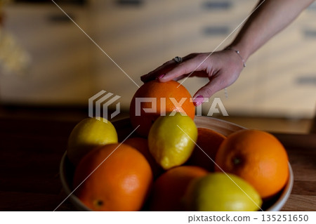 Hand reaching orange above bowl, warm lowlight kitchen, wooden table, mixed oranges and lemons, polished nails with ring, farmtotable slowliving mood, natural texture focus Hand reaching orange above bowl, warm lowlight kitchen, wooden table, mixed oranges and lemons, polished nails with ring, farmtotable slowliving mood, natural texture focus 135251650