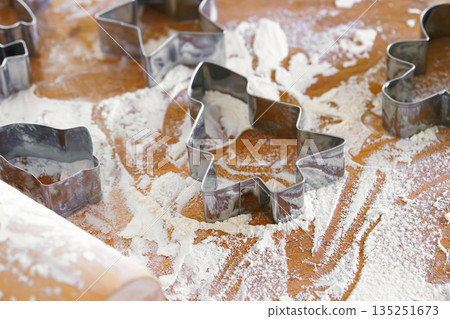 Baking gingerbread cookie cutters, wooden rolling pin, and flour are messily spread on kitchen table 135251673
