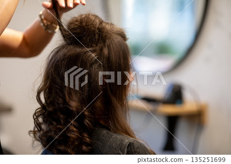 Professional creating textured curly hairstyle near mirror in salon, Hairdresser shaping and lifting top curls while client sits with cape in welllit salon environment Professional creating textured curly hairstyle near mirror in salon, Hairdresser shaping and lifting top curls while client sits with cape in welllit salon environment 135251699