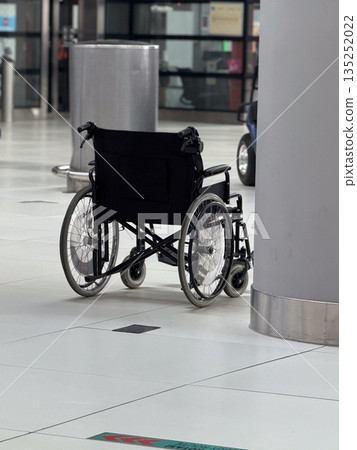 Empty wheelchair placed near column inside airport terminal hall. Mobility assistance availability, accessibility awareness, inclusive transport services, and support infrastructure concept. 135252022