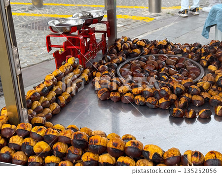 Roasted chestnuts arranged on a metal tray at a street food stand. Seasonal street cuisine, traditional roasting methods, and warm comfort food associated with autumn markets. Roasted chestnuts arranged on a metal tray at a street food stand. Seasonal street cuisine, traditional roasting methods, and warm comfort food associated with autumn markets. 135252056
