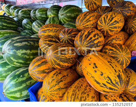 Fresh watermelons and striped melons stacked at a local market stall. Seasonal harvest abundance, food supply, agriculture diversity and natural nutrition in everyday retail environment. Fresh watermelons and striped melons stacked at a local market stall. Seasonal harvest abundance, food supply, agriculture diversity and natural nutrition in everyday retail environment. 135252057