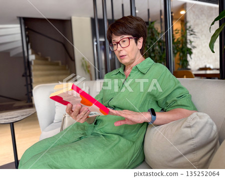 Middle-aged woman in green dress reading book on sofa. Knowledge, leisure, and personal time in modern lifestyle. Middle-aged woman in green dress reading book on sofa. Knowledge, leisure, and personal time in modern lifestyle. 135252064