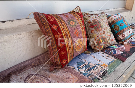 Assorted decorative pillows with traditional patterns arranged on outdoor bench. Textile craft, artisan heritage, and colorful display in cultural retail setting. Assorted decorative pillows with traditional patterns arranged on outdoor bench. Textile craft, artisan heritage, and colorful display in cultural retail setting. 135252069