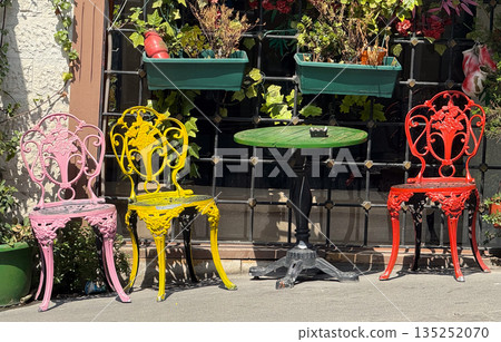 Brightly painted chairs and a green table placed outdoors near a window with plants. Decoration, leisure, and lifestyle in urban cafe atmosphere. Brightly painted chairs and a green table placed outdoors near a window with plants. Decoration, leisure, and lifestyle in urban cafe atmosphere. 135252070