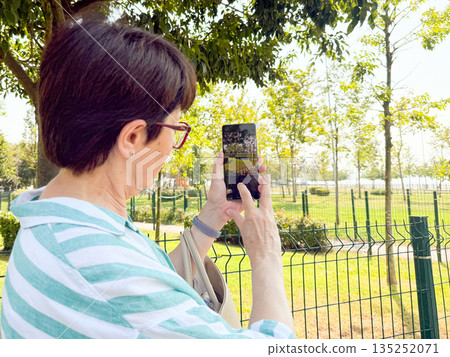 Woman taking a photo outdoors with a smartphone in her hands. Daily life, urban leisure, and personal perspective in an open park environment. Woman taking a photo outdoors with a smartphone in her hands. Daily life, urban leisure, and personal perspective in an open park environment. 135252071