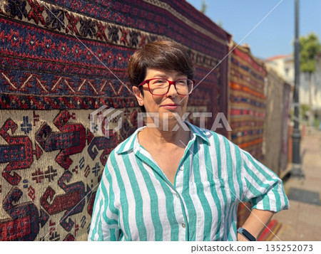 Woman standing in front of traditional patterned carpets displayed outdoors. Textile culture, personal presence, and artisan craft with heritage design and lifestyle. Woman standing in front of traditional patterned carpets displayed outdoors. Textile culture, personal presence, and artisan craft with heritage design and lifestyle. 135252073