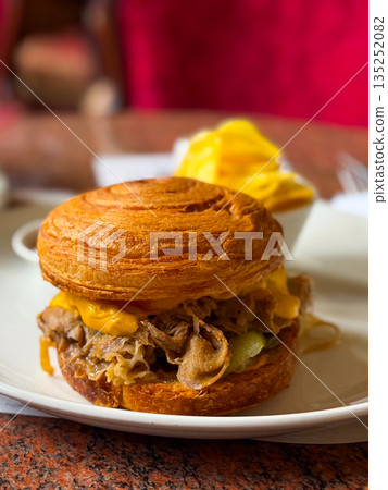Croissant sandwich with meat and melted cheese served on a plate in a cozy cafe close-up. Gastronomy, lifestyle, and casual dining experience with comfort and indulgence. Croissant sandwich with meat and melted cheese served on a plate in a cozy cafe close-up. Gastronomy, lifestyle, and casual dining experience with comfort and indulgence. 135252082