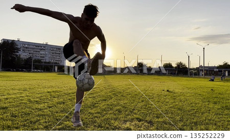 Young man juggling soccer ball on stadium at sunset. Professional footballer kicking ball at green field. Sportsman practicing tricks at meadow with sunlight at background. Freestyle football 135252229