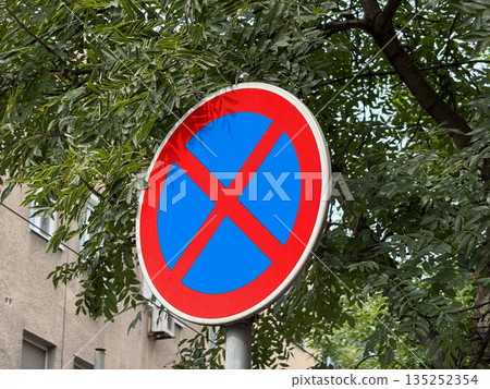 No parking road sign with red cross on a blue circle among green tree branches. Urban traffic regulation, public safety, and street infrastructure 135252354