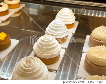 White cream swirl cakes displayed on a pastry counter under warm light. Minimal dessert design, smooth texture, and modern bakery elegance White cream swirl cakes displayed on a pastry counter under warm light. Minimal dessert design, smooth texture, and modern bakery elegance 135252368