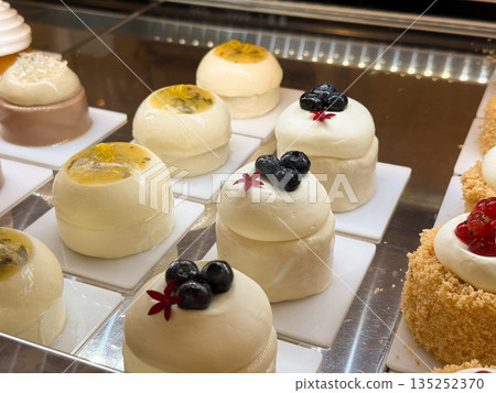 Soft mousse cakes topped with blueberry and passion fruit in a pastry case. Delicate dessert styling, fresh fruit garnish, and luxury sweets Soft mousse cakes topped with blueberry and passion fruit in a pastry case. Delicate dessert styling, fresh fruit garnish, and luxury sweets 135252370