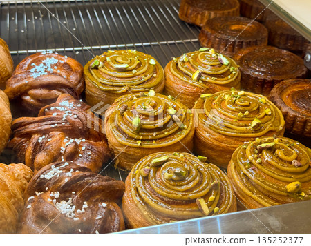 Pastry display with pistachio swirl buns and glazed bakery goods behind glass. Artisan baking, dessert variety, and cafe counter indulgence 135252377
