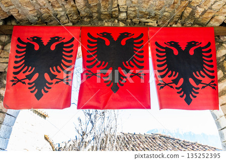 Traditional Albanian flag with black double headed eagle on red fabric hanging under stone arch, symbol of national identity, culture and heritage, Berat, Albania Traditional Albanian flag with black double headed eagle on red fabric hanging under stone arch, symbol of national identity, culture and heritage, Berat, Albania 135252395