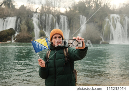 Woman taking selfie or making video while holding Bosnia and Herzegovina flag in front of Kravica Waterfall. Modern travel lifestyle with smartphone, nature scenery, winter mood, and personal memory Woman taking selfie or making video while holding Bosnia and Herzegovina flag in front of Kravica Waterfall. Modern travel lifestyle with smartphone, nature scenery, winter mood, and personal memory 135252954