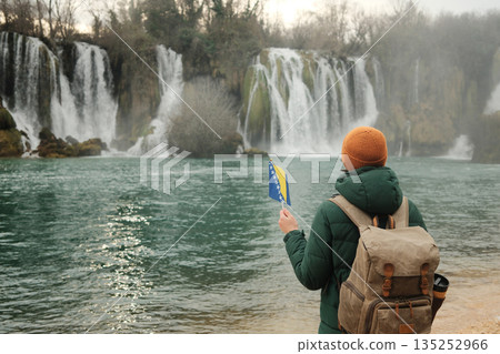 Woman with backpack holding Bosnia and Herzegovina flag while facing Kravica Waterfall. Travel reflection concept with powerful nature, winter landscape, calm water, and outdoor atmosphere Woman with backpack holding Bosnia and Herzegovina flag while facing Kravica Waterfall. Travel reflection concept with powerful nature, winter landscape, calm water, and outdoor atmosphere 135252966