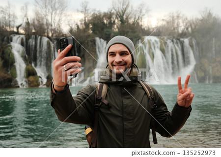 Traveler taking selfie and showing peace sign near Kravica Waterfall in Bosnia and Herzegovina. Modern travel lifestyle with smartphone, outdoor adventure, winter nature, and relaxed emotion 135252973