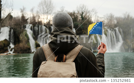 Traveler seen from behind holding Bosnia and Herzegovina flag while facing Kravica Waterfall. Exploration and reflection concept with wild nature, water scenery, and winter calm Traveler seen from behind holding Bosnia and Herzegovina flag while facing Kravica Waterfall. Exploration and reflection concept with wild nature, water scenery, and winter calm 135252981