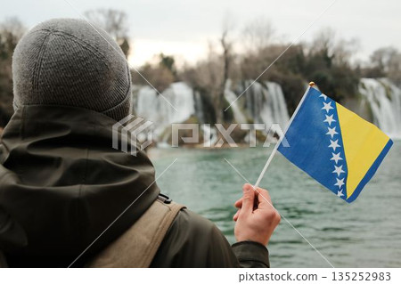 Traveler seen from behind holding Bosnia and Herzegovina flag while facing Kravica Waterfall. Exploration and reflection concept with wild nature, water scenery, and winter calm 135252983